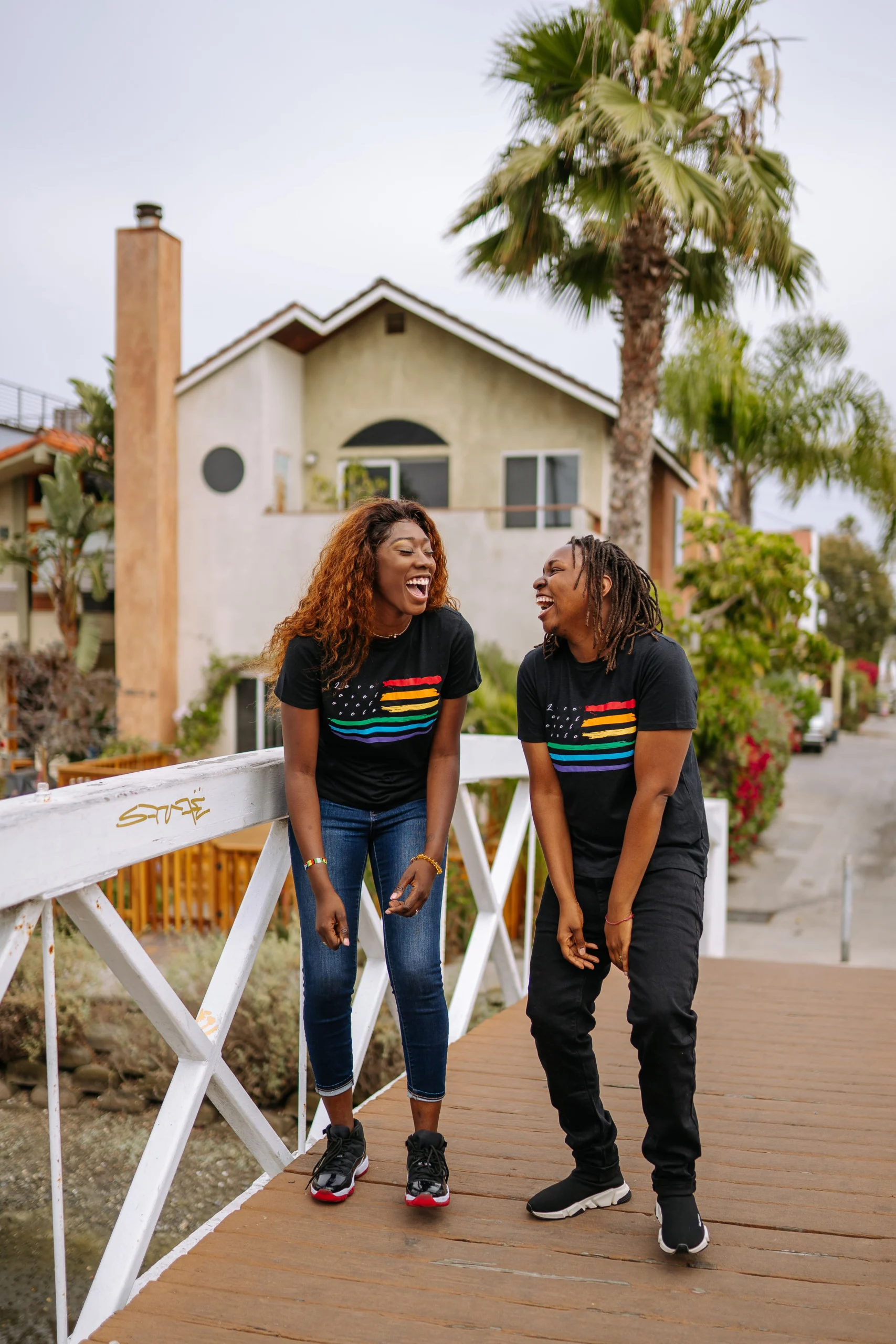 Two young Black friends laughing together on a wooden footbridge, both wearing black T-shirts printed with rainbow pride stripes.