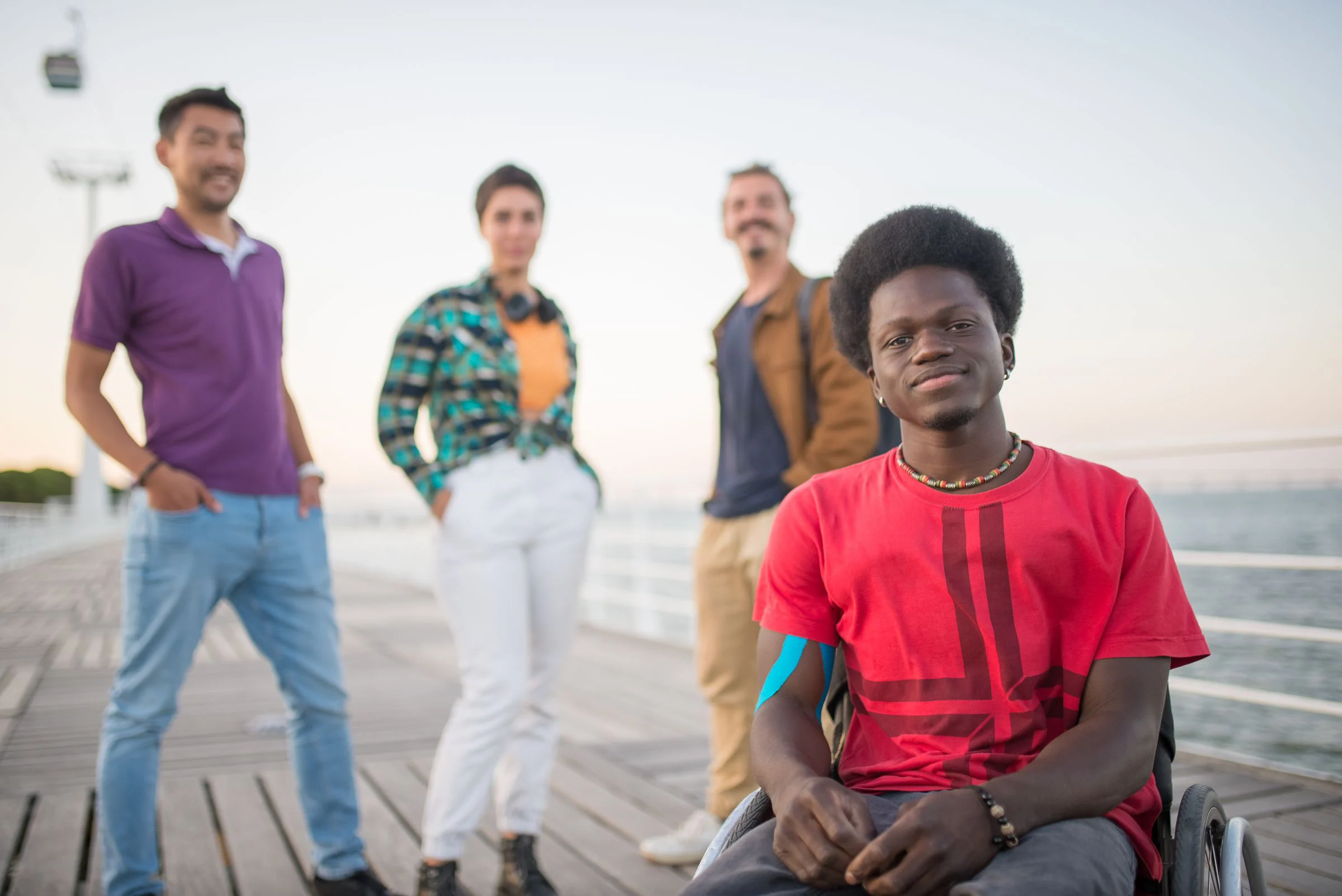 A young Black man using a wheelchair smiles at the camera on a waterfront boardwalk at dusk, with three friends standing out of focus behind him.