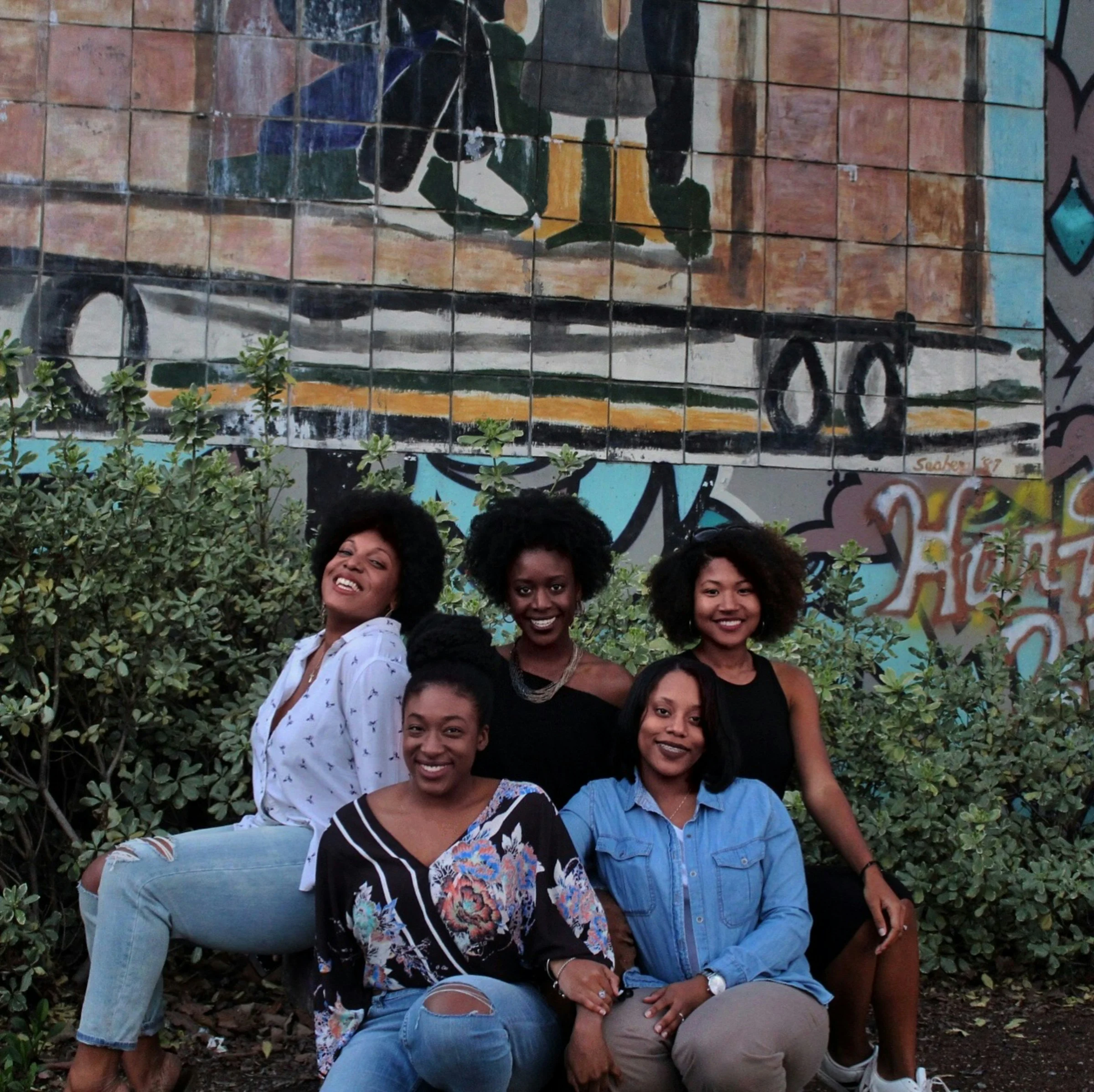 Five Black women smiling together in front of a colourful mural