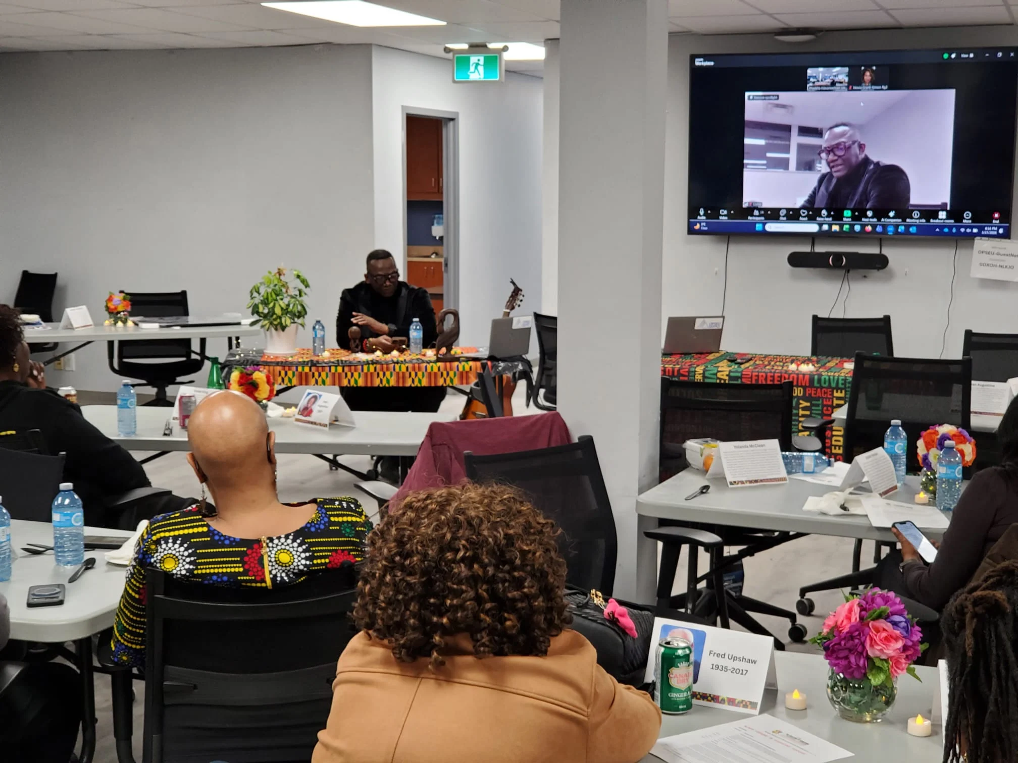 A speaker seated at a kente-draped table addresses guests, with a second speaker joining on a video screen