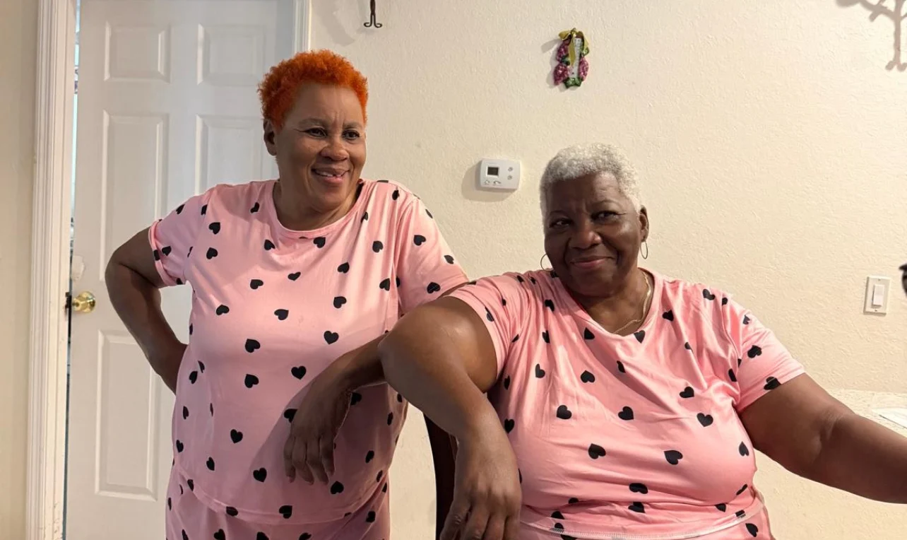 Two older women in matching pink heart-print outfits pose and smile